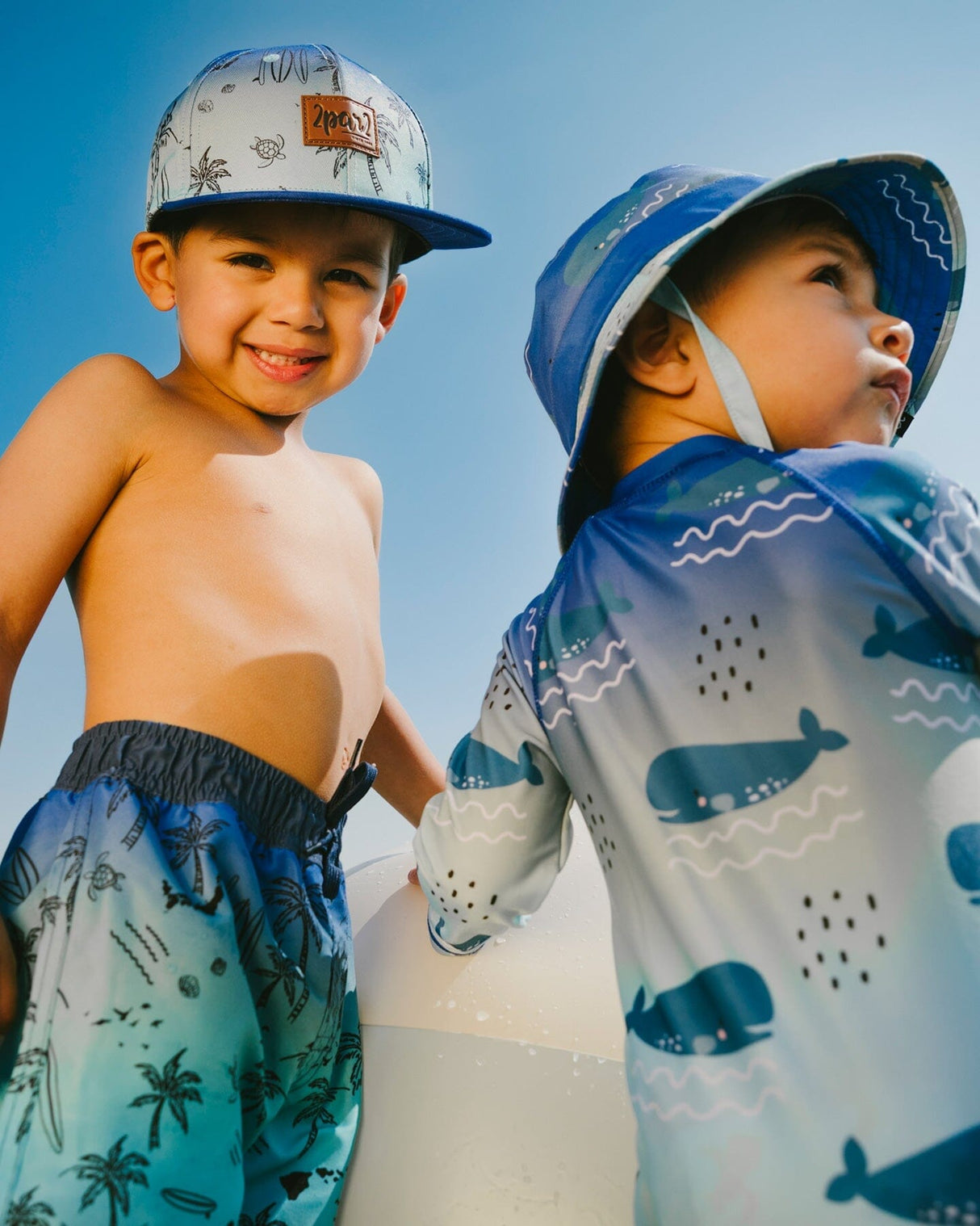Two boys wearing blue beach print boardshorts and rashguards standing by the water