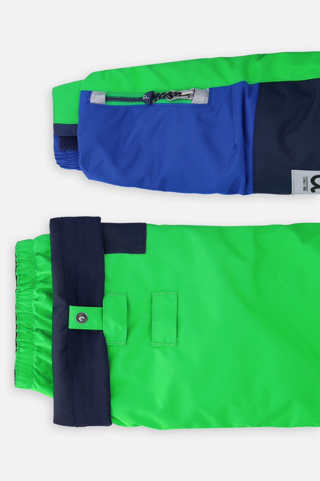 Close-up of the PLAY jacket sleeve ski-pass pocket and the adjustable green pant hem