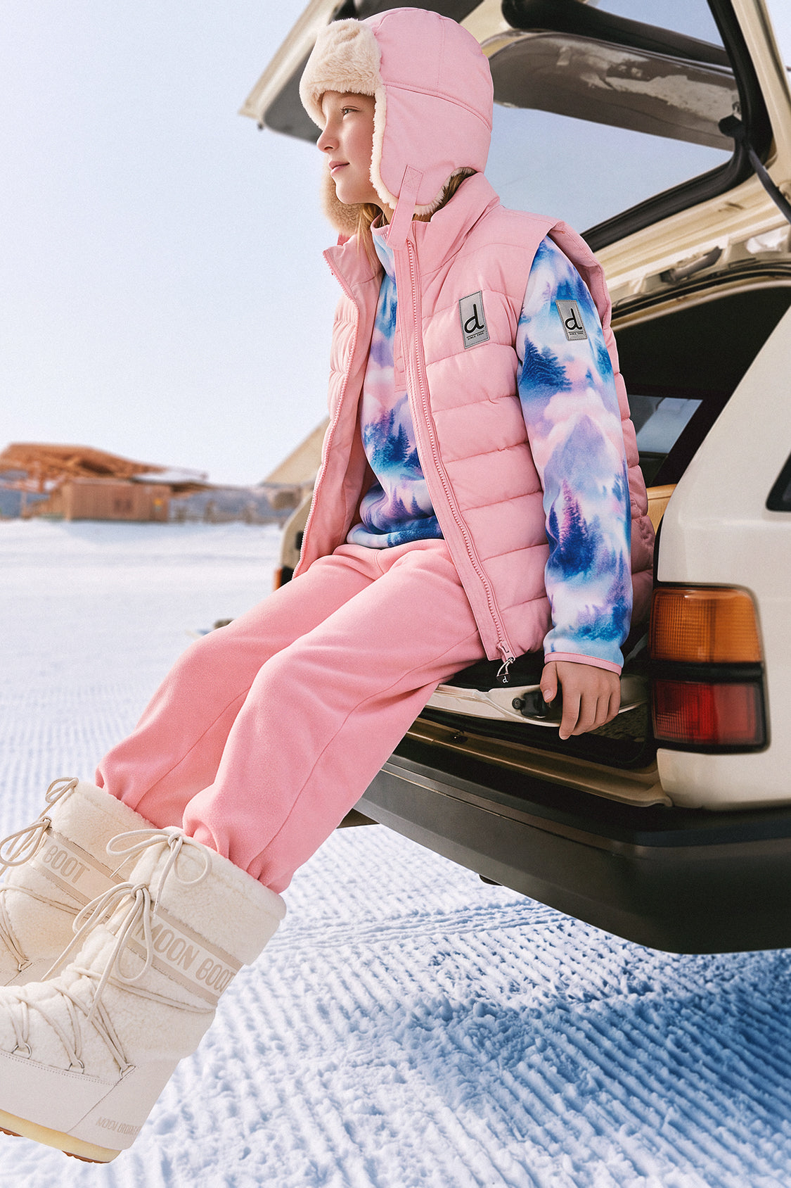 Girl sitting on a car in the snow wearing the Deux par Deux light pink lined chapka, lifestyle photo