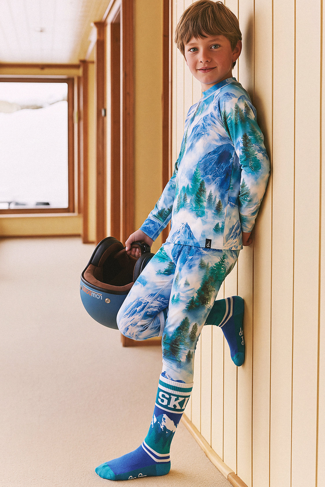 Boy leaning on a wall in the Deux par Deux two-piece thermal underwear set in blue mountain print, holding a ski helmet
