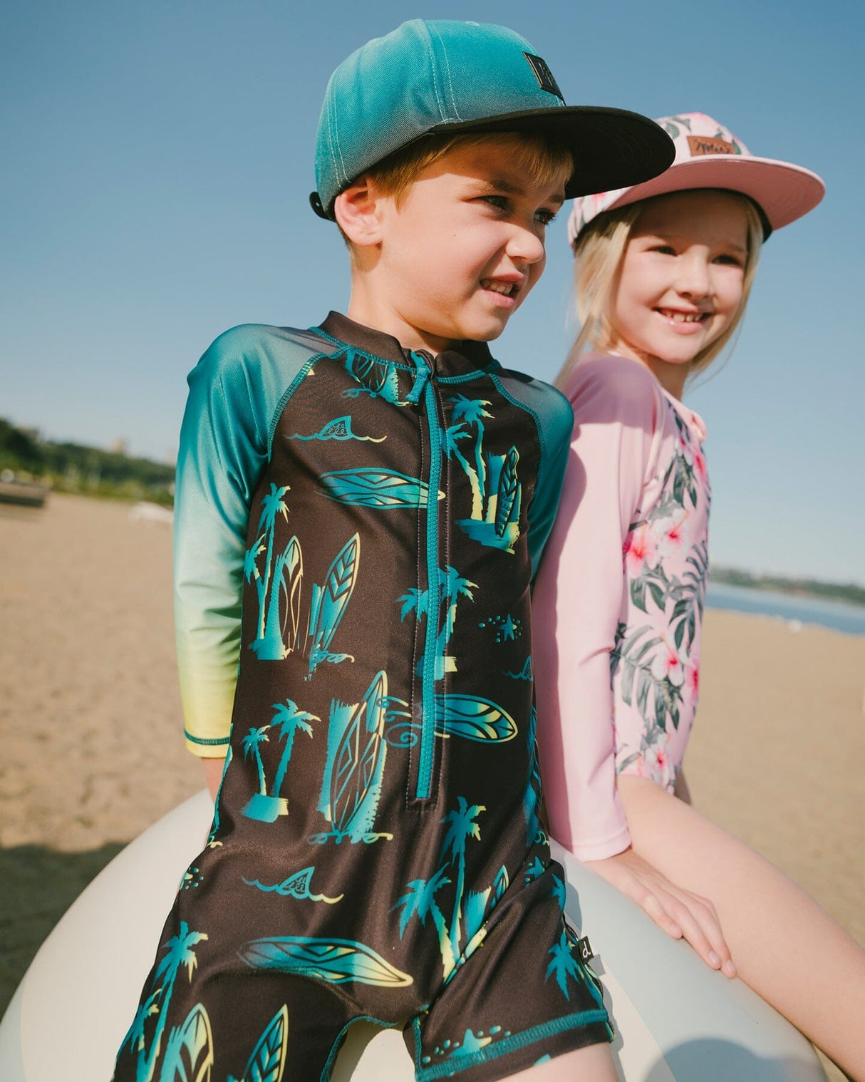 Two boys on the beach wearing surf print one-piece rashguard suits and matching hats