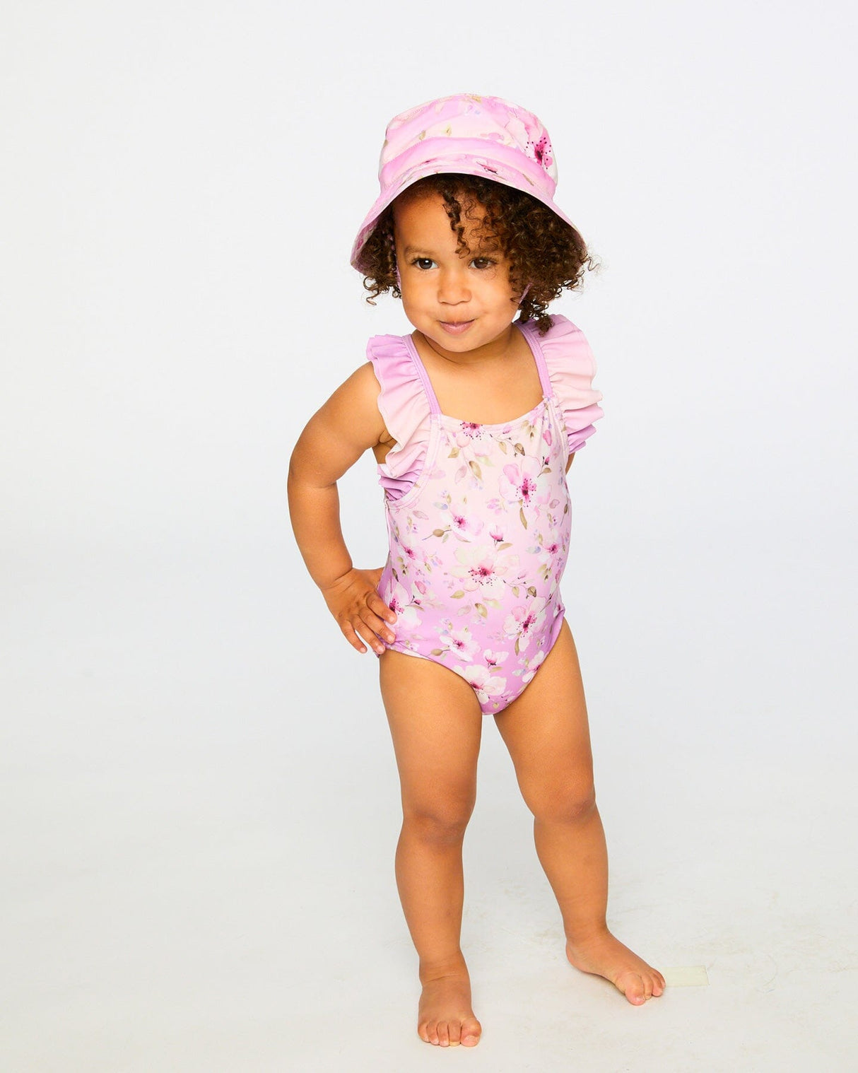 Toddler girl wearing purple floral beach hat and matching swimsuit front view