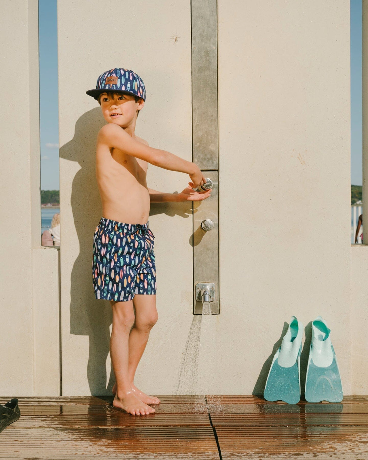 Boy leaning on dock railing wearing blue surfboard boys swim trunks front view