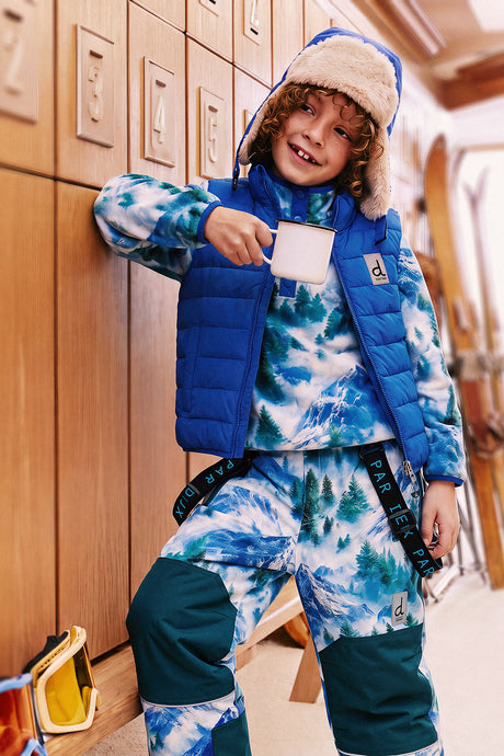 Boy standing in a ski lodge wearing the Deux par Deux blue lined chapka with a matching snow outfit