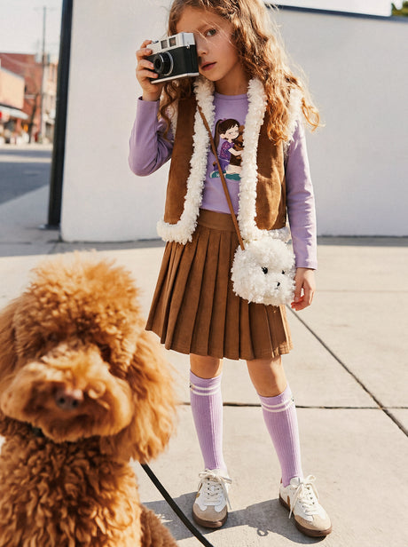 Child wearing the reversible vest, brown side out, with a pleated skirt and a fluffy bag while holding a camera outdoors