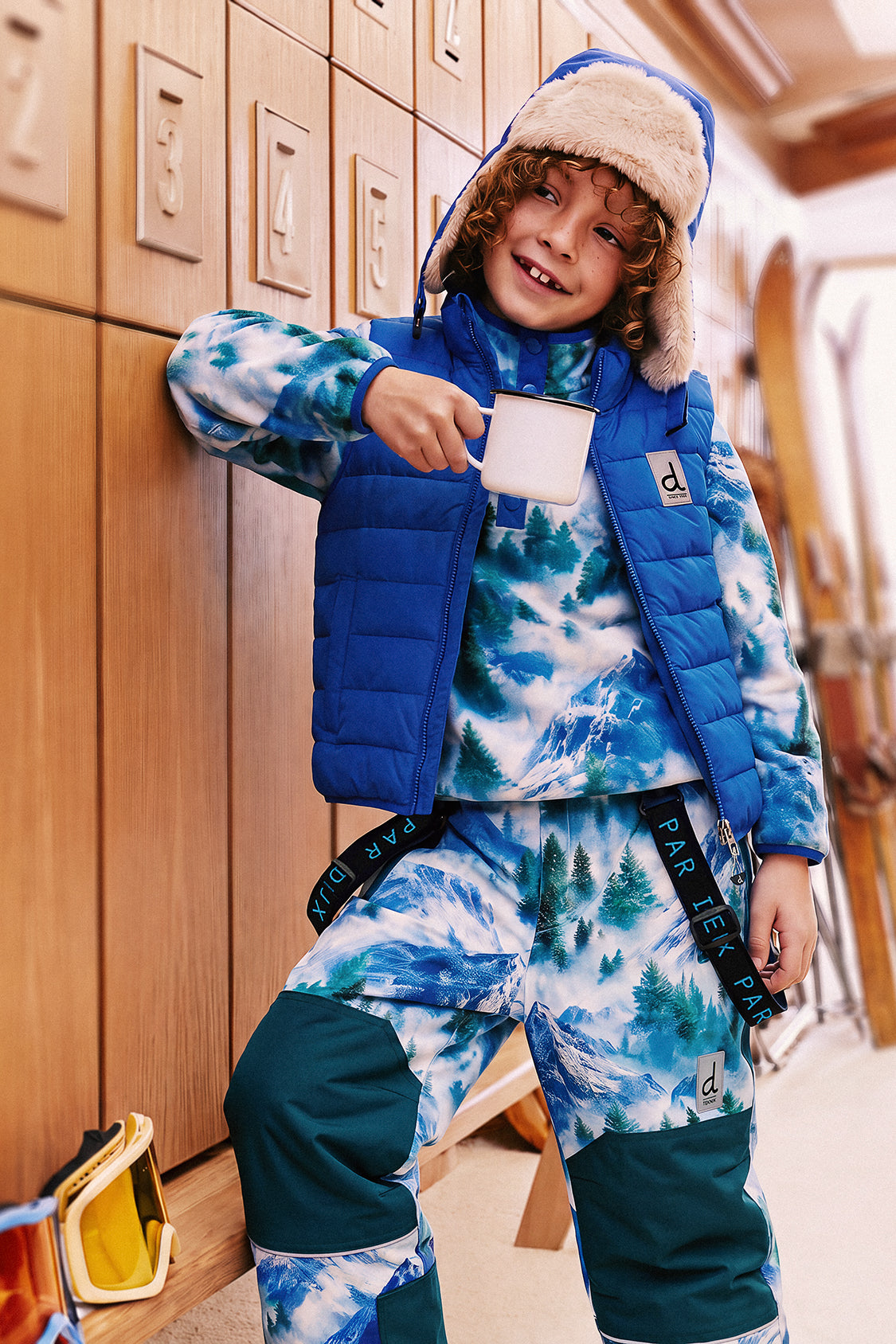 Smiling child by wooden ski lockers wearing the blue Deux par Deux quilted vest and a trapper hat, holding a mug