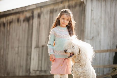 Little girl wearing pastel knit sweater and pink corduroy skirt, standing with fluffy white poodle – stylish fall kids outfit from Jenni Kidz Canada.