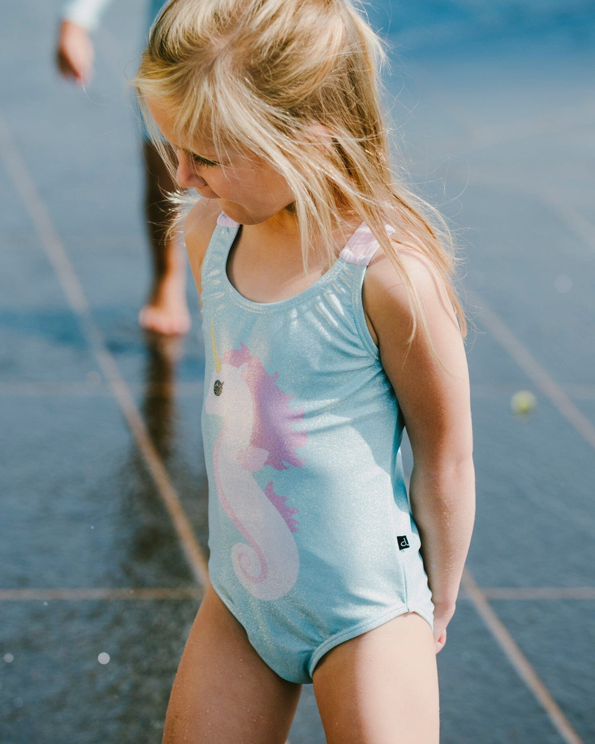 Girl standing by the water wearing aqua seahorse swimsuit side view
