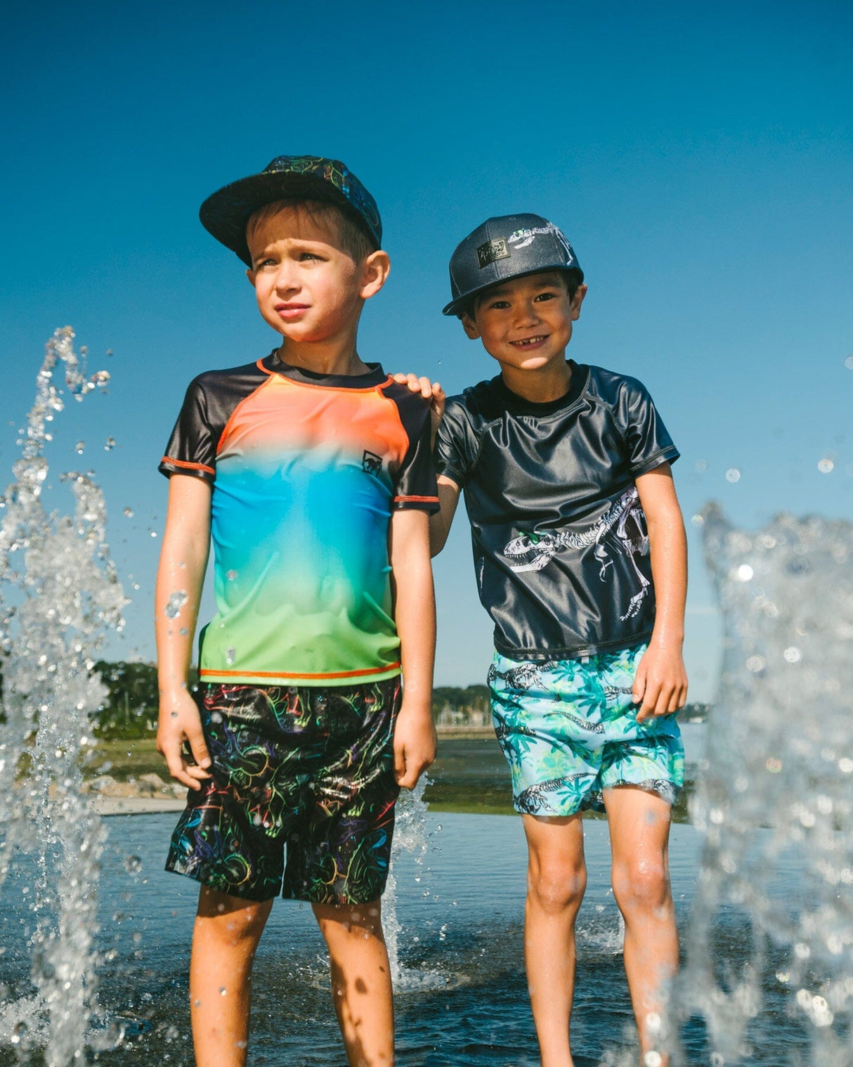 Two boys standing in matching swim sets with black dinosaur rashguard tops by the water