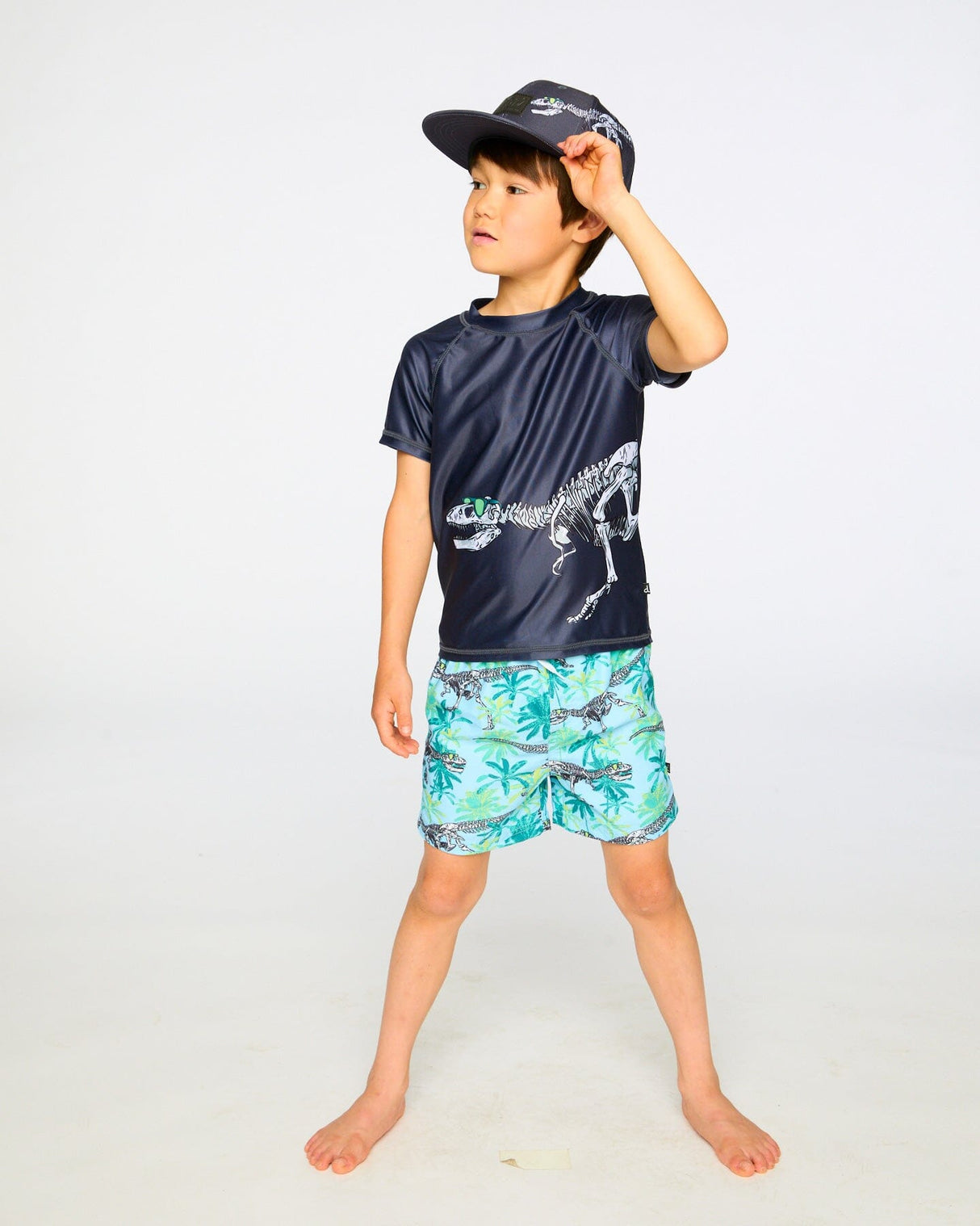 Boy in studio wearing black dinosaur rashguard and printed swim trunks front view