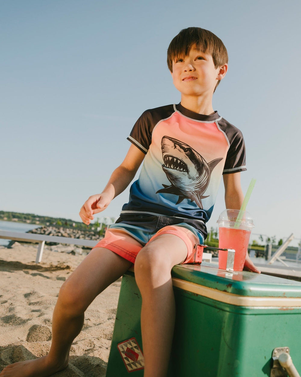 Boy sitting near the water wearing blue/orange gradient shark rashguard and matching trunks