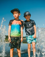 Two boys standing on the beach wearing black dinosaur swim trunks and rashguards