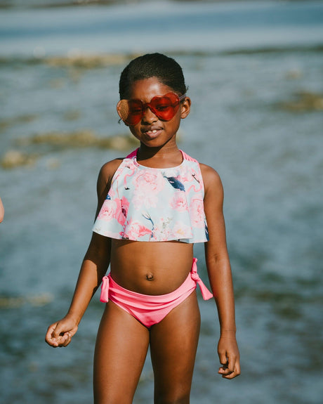 Girl standing by the water wearing blue flamingo ruffle bikini top and pink tie-side bottoms