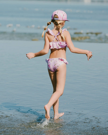 Girl walking along the shoreline in purple flower two-piece swimsuit and sun hat side view
