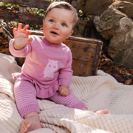 Smiling baby girl in a Deux par Deux pink knit outfit waving while sitting on a blanket outdoors