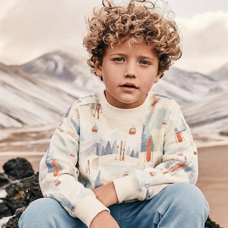 Boy in a Deux par Deux beige ski print fleece sweatshirt sitting outdoors in the mountains
